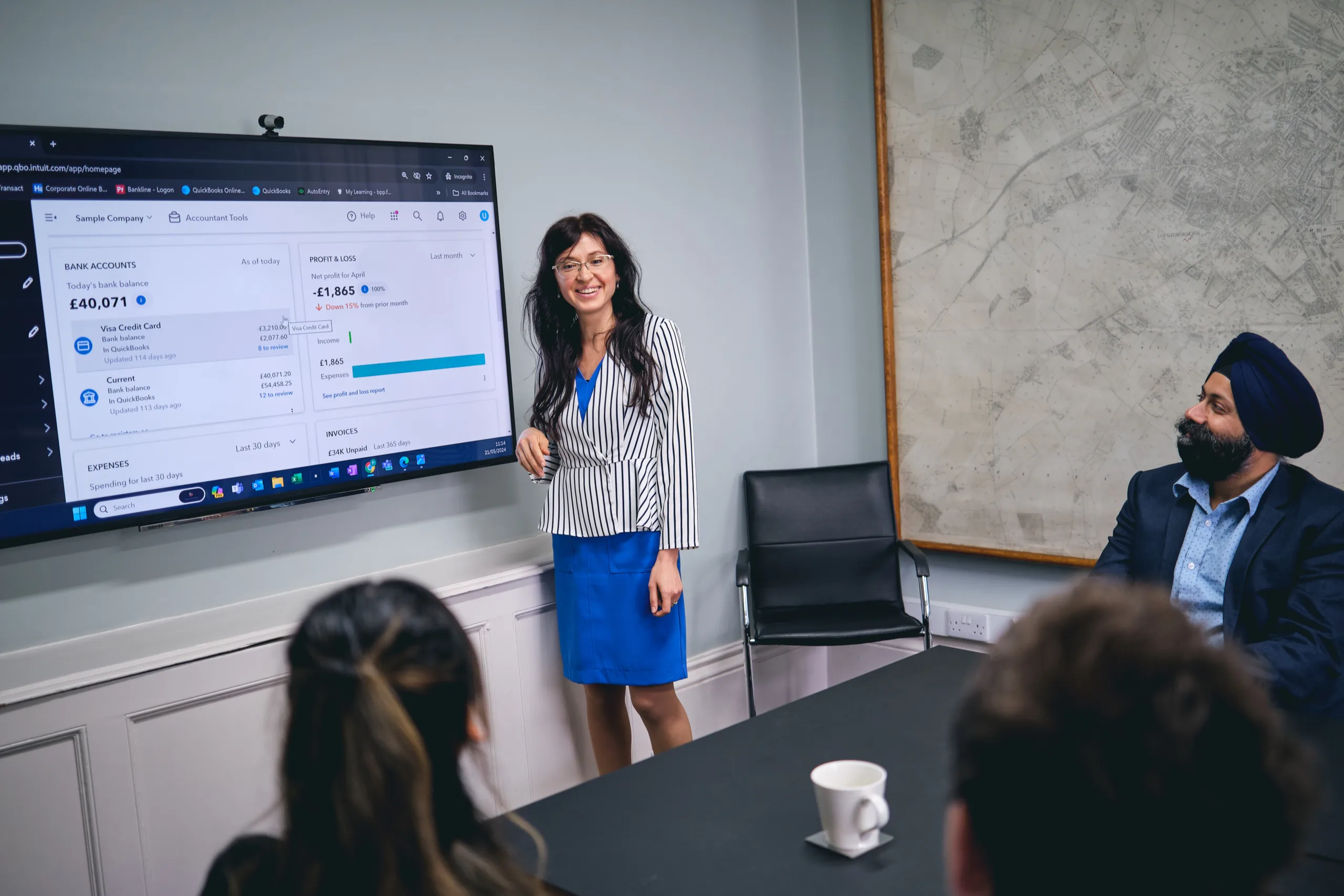 Decorative image: woman in bright blue skirt stands next to TV screen with accounting data, presenting to 3 people sat at a table in front of her. To accompany blog on accounting for women in business.