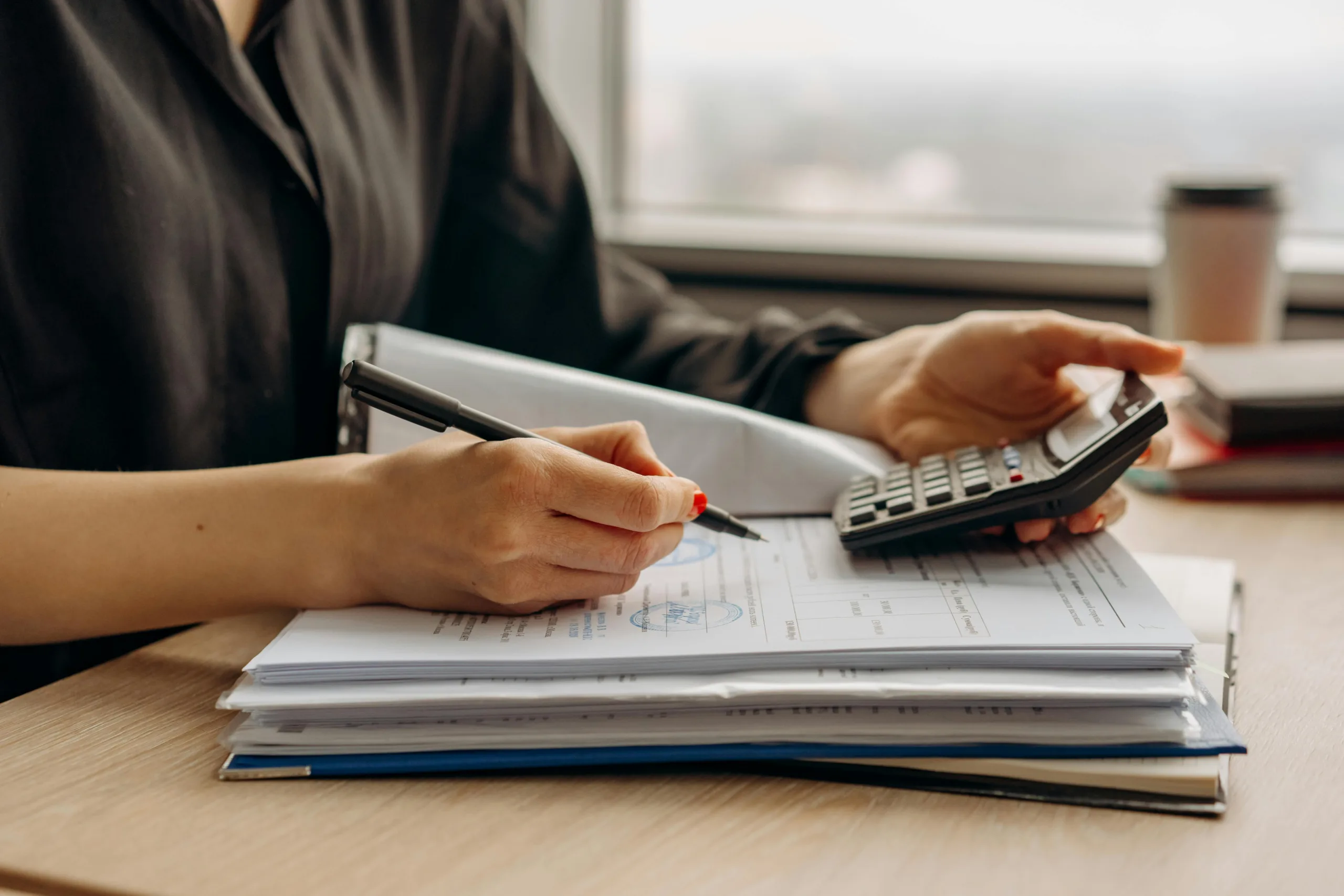 Decorative image to accompany blog on budget tips for new business startups. Close up shot of a woman with painted red nails holding a calculator in her left hand and writing on forms with the pen in her right hand.