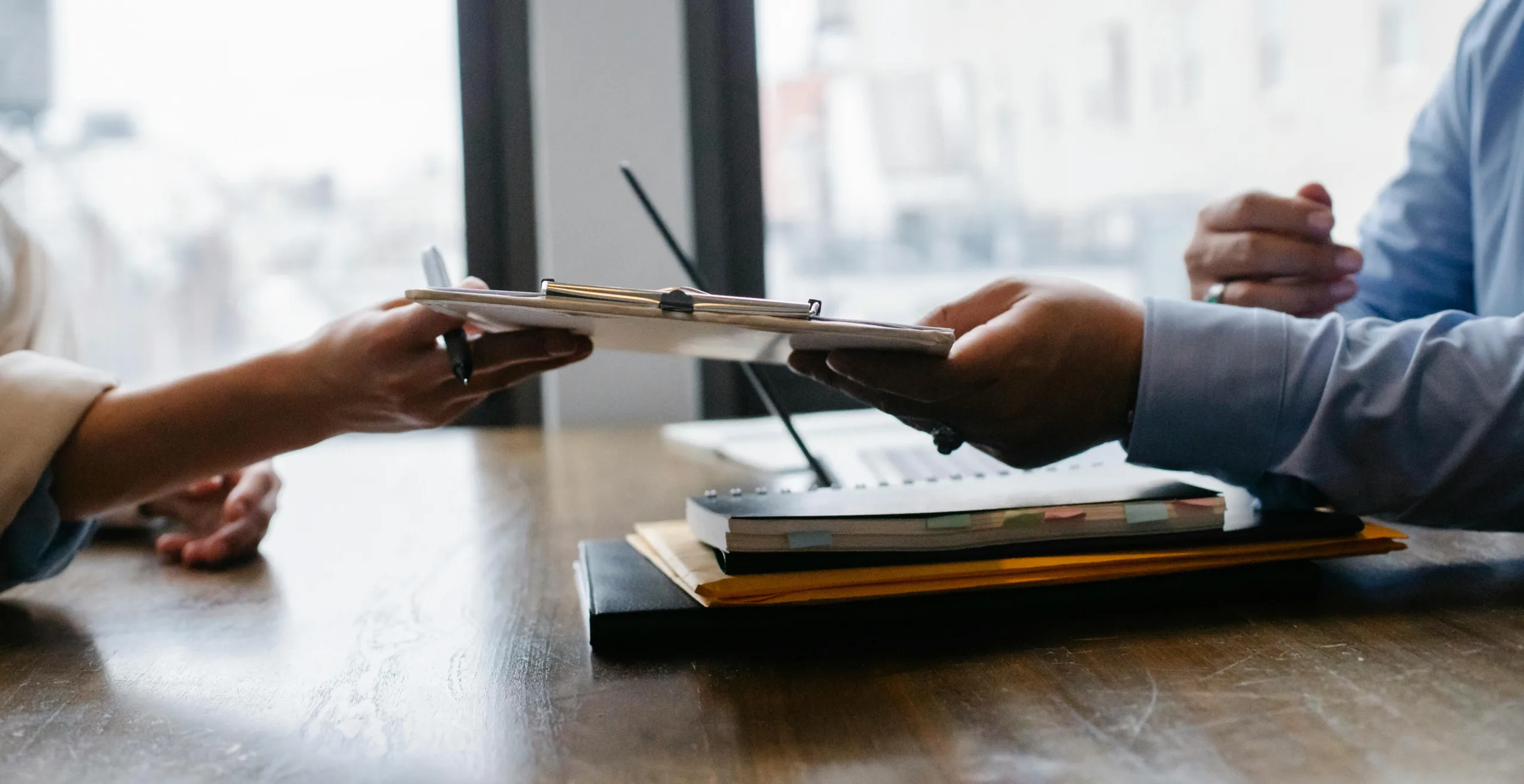 Decorative image to accompany blog on the benefits of outsourcing your accounting: close up image of a desk with papers on and hands passing papers from one to the other
